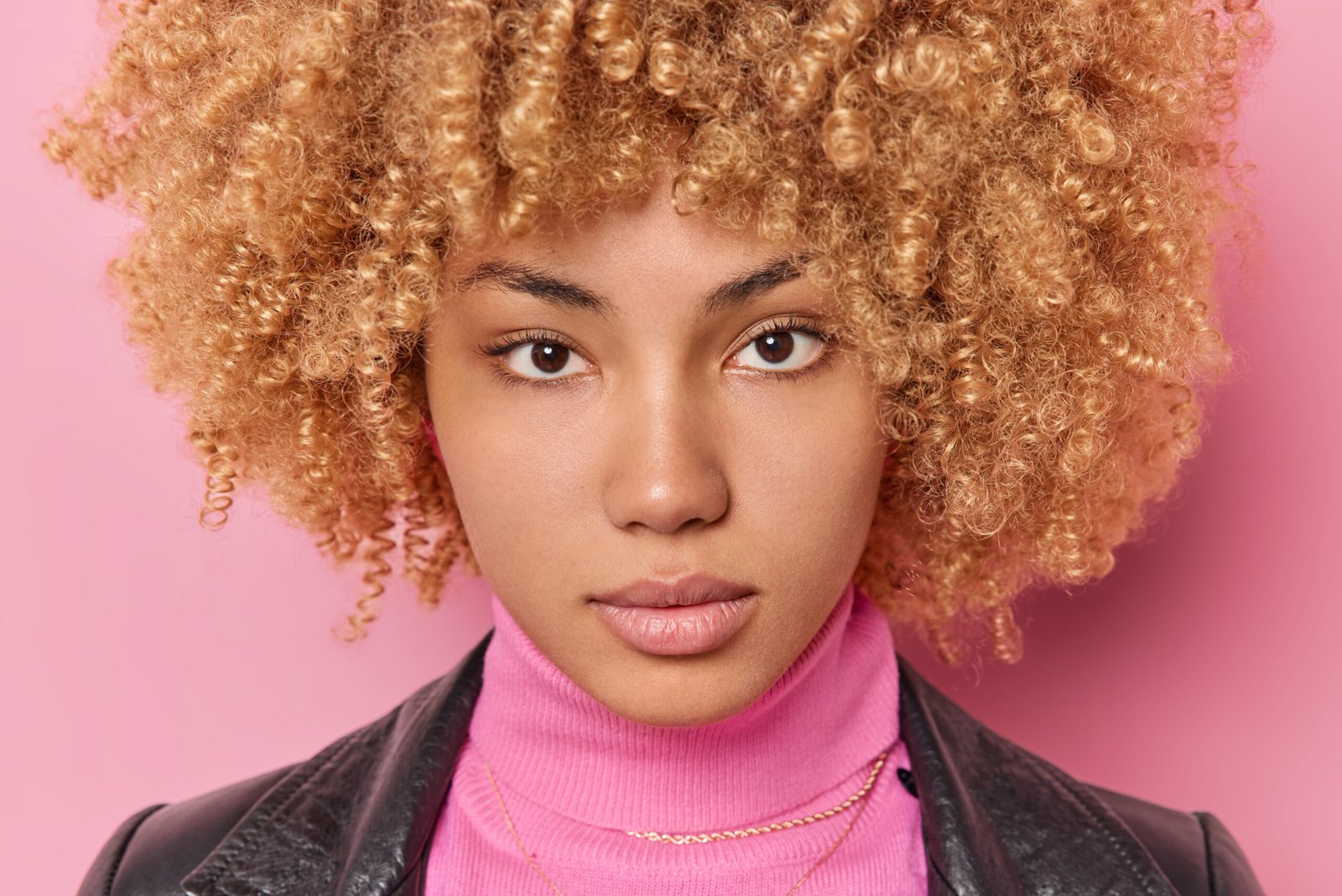 Close up portrait of serious woman looks self confident directly at camera has healthy skin curly bushy hair wears turtleneck and leathes jacket isolated over pink background. Human face expressions